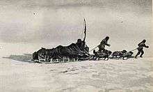 A loaded sledge being pulled across an icy surface by two figures and a team of dogs