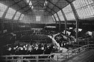 Darkened interior of a large hall with two rows of high windows along each side. It is possible to discern a seated orchestra in the foreground, with mass choirs in the background.