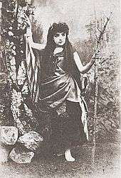 Full shot of Bond on stage with rustic scenery and holding a staff.