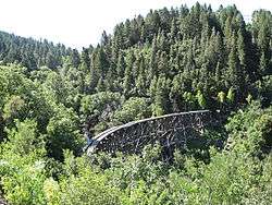 A broken trackless trestle rises up and over the green treetops of a small mountain canyon.