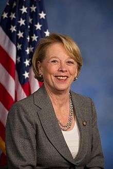 A photograph of a middle-aged woman with short, blonde hair, smiling.  She wears a gray suit, and is posed sitting before an American flag, hanging behind her to the left.