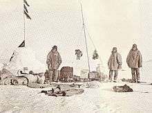 On the left is a snow cairn with flags. Three men are nearby, and assorted equipment is strewn on the snow.