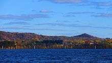 A landscape photo of Onalaska Wisconsin taken in the fall from Lake Onalaka shows a sunny sky of blue above with clouds and the cool blue waters of Lake Onalaska below in the foreground. Sandwiched between is a horizontal ribbon that shows the city elevated 100 feet above the water and tree covered bluffs in the not to distant background. A few houses along highway 35 are barely visible.