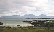 A small sandy beach lies beyond a grassy foreshore. In the distance the outline of a range of brown and grey hills is visible under cloudy skies.