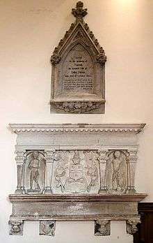 Two elaborate stone tablets set into a plain wall and lit from above.  One is pentagonal and has black writing in an arched recess; the other, immediately below, is larger, rectangular and has effigies of three people.