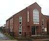 A modern building in two shades of red brick, behind a low black fence. White-framed three-paned windows line the side wall and flank the entrance—a double door sheltered below a protruding section supported on brick columns. This has a plaque reading "QUEEN STREET CHURCH CENTRE" and a pentagonal window.