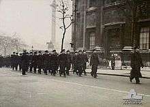 Black-and-white photo of a large group of men wearing military uniforms marching in formation along a city street