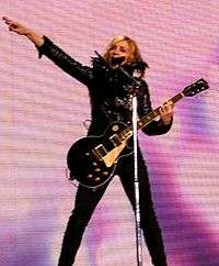 A woman in black clothing holding a guitar and standing behind a microphone stand with one arm extended straight into the air. In the background is a screen with shades of pink and purple.