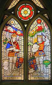 Two men in armour stand opposite each other. They wear crowns and hold swords in their hands. Above the man on the left is a flag of a white boar and a white rose. Above  the man on the right is a flag of a red dragon and a red rose. Above and between the two roses is a white rose superimposed on a red rose.