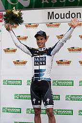 A man in his early twenties wearing a white and black cycling jersey with blue trim and a matching cap. He is holding his arms up in a posture of victory, and holds a bouquet in his right hand.