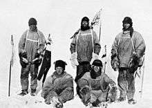 Five men(three standing, two sitting on the icy ground) in heavy polar clothing. All look unhappy. The standing men are carrying flagstaffs and a Union flag flies from a mast in the background. Scott's party at the South Pole. Left to right: Oates; Bowers; Scott; Wilson; Evans