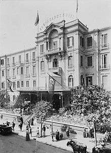 Period photo of a hotel, with four stories, large square windows, and a wrought iron portico with flags. Pedestrians, horse-drawn carriages and a motor car are before it