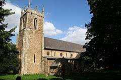 Stone church with a square tower. Unusually the windows in the nave are circular. The tower is on the left and dominates the picture. It has a crenellated flat top with stone pinnacles at each corner, narrow arched-top bell ports high up and very narrow slit windows lower down. The nave is receding left to right and is partly obscured by one of the two large yews that frame the picture. In shadow the porch can just be made out near the tower. The day is sunny and the sky mostly blue. The stonework of the church appears golden, similar to Cotswold stone. In the short grass of the foreground the top of a single gravestone can be seen.