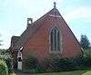 A red-brick church with a tiled roof extending nearly to ground level. Low shrubs surround the building on all sides. A three-light lancet window with stone mullions dominates the nearest side. A small stone cross and bell-tower are on the roof. The left-hand side has a white entrance porch.