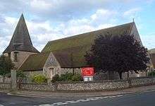 Three-quarter view of a long flint church with an extremely wide, stumpy tower on the left. This is topped with a dumpy grey spire. The brown church roof is heavily discoloured. A tree in full leaf obscures the near corner.
