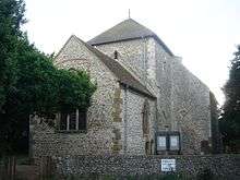 A flint rubble church with large stone quoins, behind a low flint wall. A three-light lancet window in the nearest wall is partly obscured by a tree in full leaf. A squat tower with a squared-off pyramidal roof stands in the middle. To its right is a blank-walled protrusion with a blocked entrance.