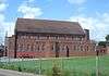 Side view of a tall, plain red brick church with an entrance porch at the left end and several uneven projections from the near side. The porch has a stone statue and an arched entrance door. The tiled roof has stone ends, both of which have small stone crosses. The lower level of the longer side has 15 unevenly spaced but identical round-headed windows; above this and immediately below the roofline are another eight windows of the same type, which are evenly spaced.