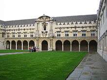 Facade of an ancient building, facing an expanse of grass. At ground floor level the building presents a long colonnade of arches, above which are rows of windows up to a crenellated roof line.