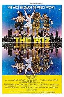 Four characters from the film dancing on top of a logo "THE WIZ". A city s以除kyline just after dusk is behind them, and the entire scene is mirrored in water before them. The people are Dorothy, the Scarecrow, the Tin Woodman, and the Lion.