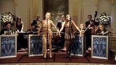 Two female vocalists perform on stage foreground to eight bandmates on the set of Gossip Girl.
