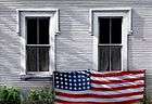 oil painting of a white clapboard house,two windows with flag draped along bottom sills, weeds along base