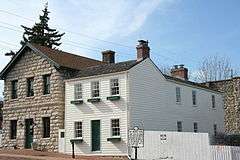 A small, two-story house with white siding panels stands between a two-story, beige brick house and a white fence.