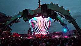 A concert stage; four large legs curve up above the stage and hold a video screen which is extended down to the band. The legs were lit up in green. The video screen has multi-coloured lights flashing on it. The audience surrounds the stage on all sides.