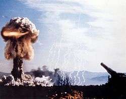 An artillery piece with a mushroom cloud rising in the distance