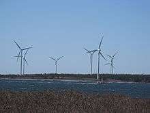 Seven three-blade wind turbines on the shore. A fishing boat is passing by. 